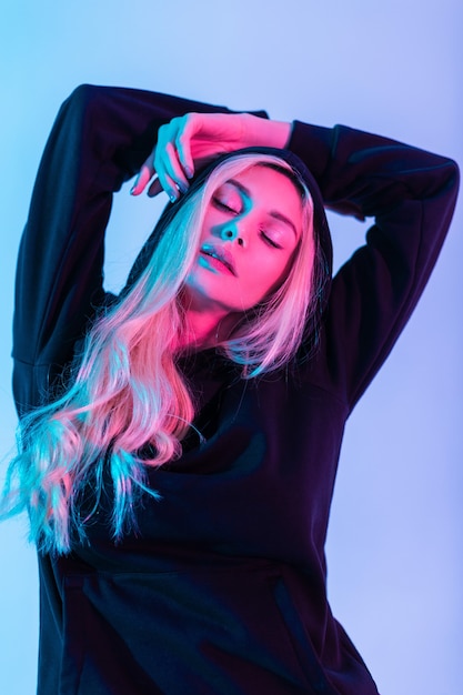 Creative color portrait of a young beautiful blonde woman with cute face in a fashion black sweatshirt wearing a hood on a multicolored pink-blue background