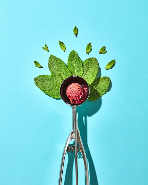 Creative composition from litchi fruit with mint leaves in the metal spoon for ice cream on a blue glass background with shadows. Food modern style, top view.