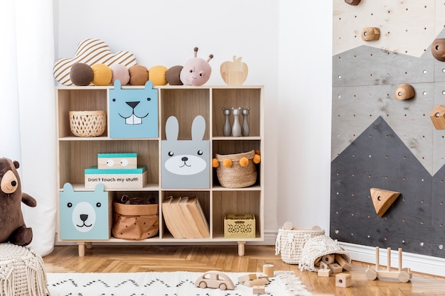 Creative composition of stylish and cozy scandi child room interior with graphic on the wall toys and plush toys sofa commode armchair carpet and accessories White walls and parquet floor