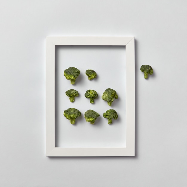 Creative composition with freshly picked green broccoli in a frame and one part out of it on a light gray wall, place for text. Flat lay. Vegan concept.