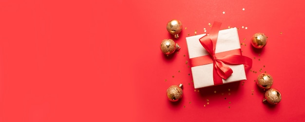 Creative composition with red present box, ribbons, red gold big and small balls, holiday decorations on red background.