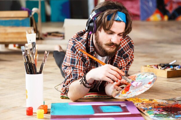 Creative darkhaired bearded hipster male artist in headphones and blue bandana lies on the floor and mixed colors on palette in his contemporary studio with many paintings