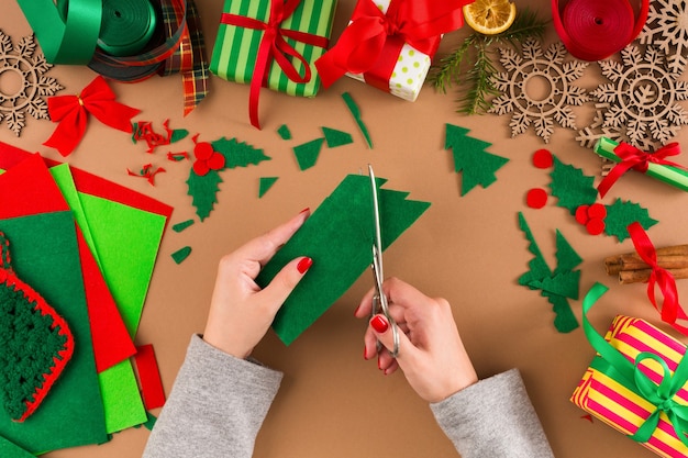 Creative diy craft hobby. woman makes felt christmas tree decoration on table with felt sheets and scraps, scissors and trappings. closeup of female hands at craft paper background