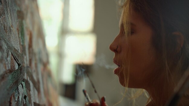 실내에서 추상적인 그림에 담배 연기를 불고 있는 창의적인 여성 예술가 직장에서 팔레트 나이프를 손에 들고 예술 작품을 보고 있는 영감을 받은 여성 미술 스튜디오에서 아침을 보내는 숙련된 화가