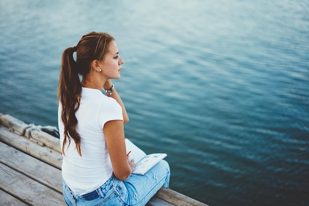 Creative female writer resting on pier near lake during making notesin in notepad for next article