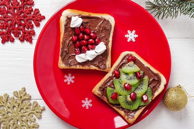 Panini festivi creativi a forma di albero di natale e cappello di babbo natale, realizzati con frutta fresca e marshmallow su un piatto rosso su uno sfondo di legno bianco. concetto di colazione per bambini.