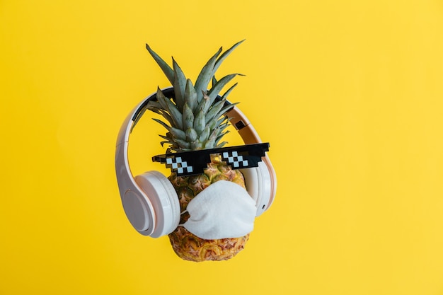 Creative funny pineapple face wearing glasses headphones and protective medical mask. Resting levitating pineapple face on color yellow summer background. Coronavirus travel concept.