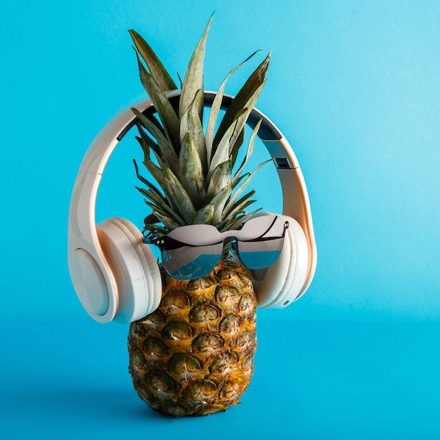 Photo creative funny pineapple face wearing sunglasses headphones. pineapple face levitating listening to music on color blue summer background. square high quality stock photo.