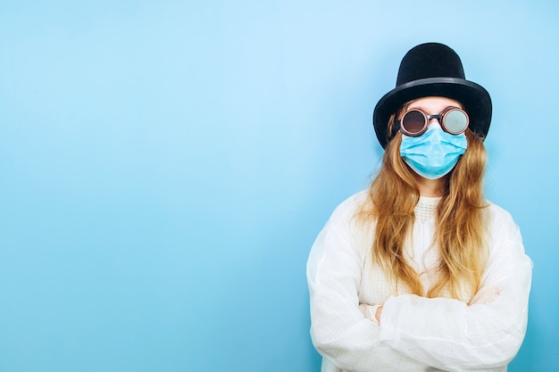 Creative girl in a white robe, medical mask, glasses and a hat on a blue background. Self-isolation during coronavirus quarantine. Epidemic covid-2019.