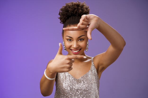 Creative good-looking african woman in silver glittering dress\
searching inspiration make finger frame look camera pleased smiling\
satisfied found perfect photo angle, standing blue background.