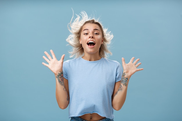 Creative happy and playful beautiful blond girl with tattoos on arms raising palms up opening mouth
