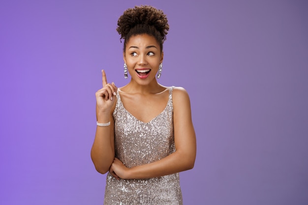 Creative joyful dreamy young african-american girl in silver\
dress raise index finger eureka gesture look aside inspired have\
awesome idea sharing suggestion tell plan aloud, standing blue\
background.