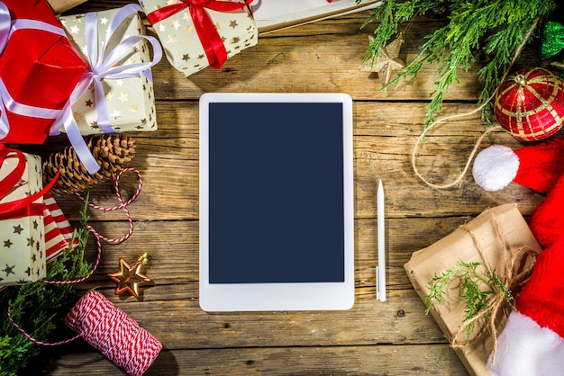 Creative layout frame with lot gift boxes, christmas tree branches, pine cones, hot chocolate mug, xmas sweet, tablet pc on old wooden background. Xmas and New Year preparation background