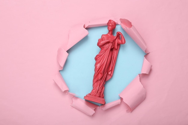 Creative layout pink antique greek goddess statue through the torn hole of pink blue paper minimalism concept art flat lay top view