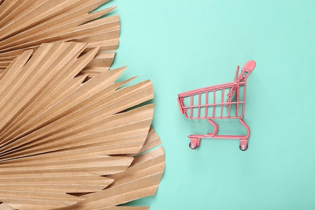 Creative layout Pink supermarket trolley on blue background with dry palm leaves Flat lay Top view
