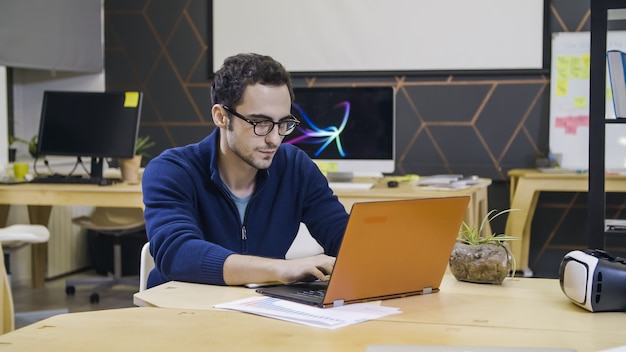 Creative man in glasses using laptop at workplace in bright modern office