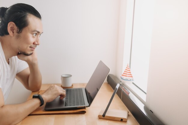 L'uomo creativo lavora con il laptop sull'area di lavoro della scrivania in legno vicino alle finestre