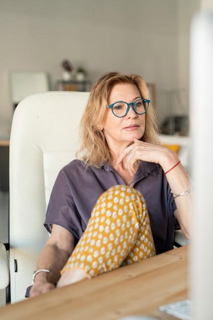 Creative mature businesswoman or web designer looking at computer screen while sitting in armchair in home environment