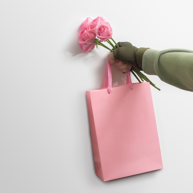 Creative men's greeting concept. A man's military hand holds roses and a gift bag leaning against a white wall.