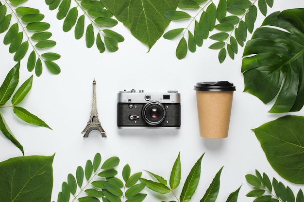 パリへの創造的なミニマルな背景旅行。緑の葉と白い背景の上のレトロなカメラ、カップ、エッフェル塔の置物