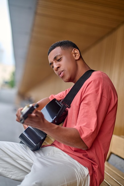 Creative mode. Young adult african american man in red t-shirt and white trousers sitting sideways to camera playing guitar enthusiastically outdoors on fine day