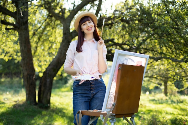 Creative painting, art, relaxation and inspiration. Pretty young female artist painter painting at sunset in beautiful spring green park or garden. Young woman holding brush for painting on canvas.