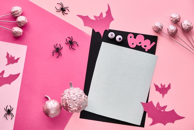 Creative paper craft Halloween flat lay in pink, light purple and black. Top view with copy-space on stack of cards, bats, chocolate eyes and decorative pumpkins.