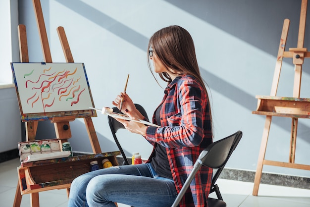 Creative pensive painter girl paints a colorful picture on canvas with oil colors in workshop.