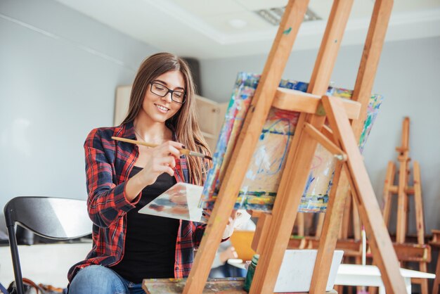 Creative pensive painter girl paints a colorful picture on canvas with oil colors in workshop.