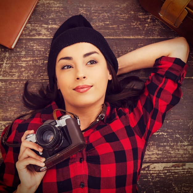 Photo creative photographer. top view of beautiful young woman in headwear lying on the floor and holding camera while suitcase and note pad laying near her