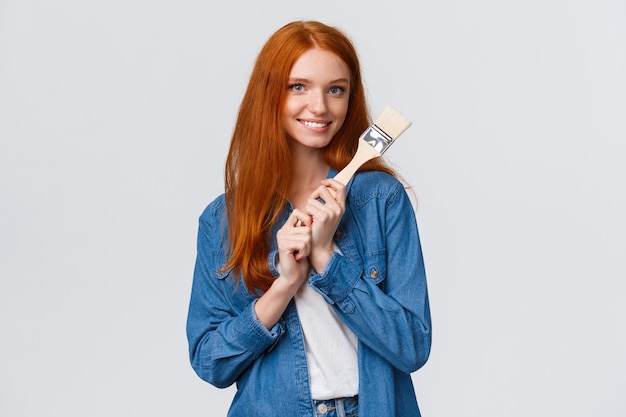 Creative pretty redhead female in denim shirt, creating new art, painting, making repair works in own room, holding paintbrush and smiling tempting, standing white 