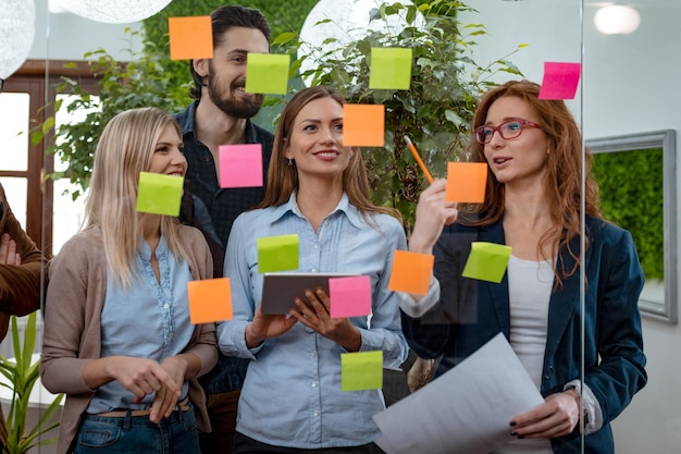 Creative professionals standing and discussing at the office behind glass wall with sticky notes and looking a post it note wall.