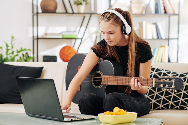 Creative serious teenage girl in wireless headphones playing guitar and writing new song on laptop