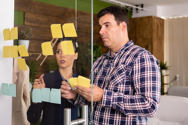 Creative team is developing a new project.Team leader puts sticky notes on glass board.