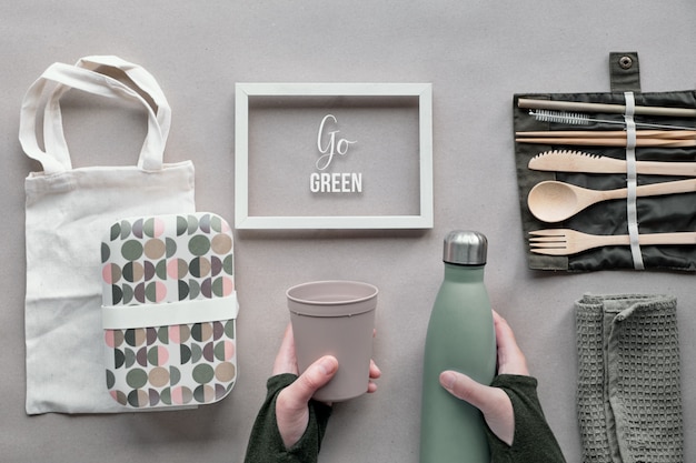 Creative top view, zero waste packed lunch concept. Flat lay, takeaway lunch set - bamboo cutlery, box, cotton bag and hand with coffee-to-go cup on brown paper. Frame with text "Go green".