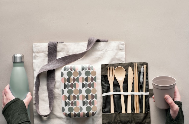 Creative top view, zero waste packed lunch concept, takeaway lunch box set with bamboo cutlery, reusable box, cotton bag and hand with coffee-to-go cup above on craft paper. Sustainable lifestyle.