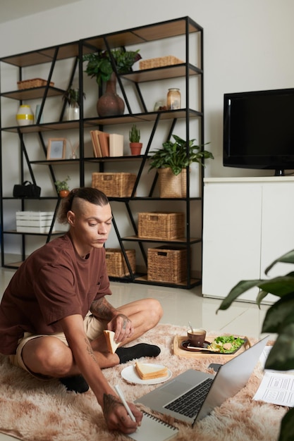 Creative ux designer sitting on the floor in his apartment eating breakfast wrting down to do list t