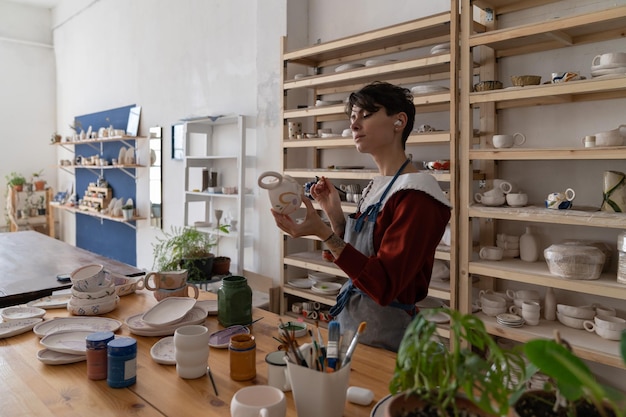 Creative woman ceramist paints handmade pottery from natural claystanding next to table in workshop