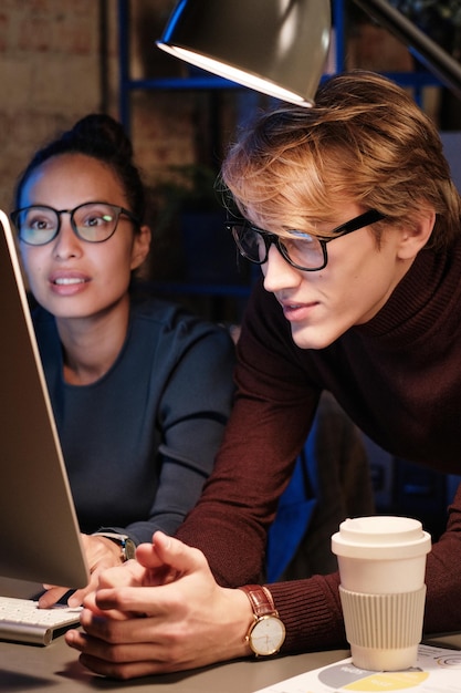 Creative young multiethnic colleagues in eyeglasses sitting in\
front of computer under lamp light and brainstorming about project\
at night