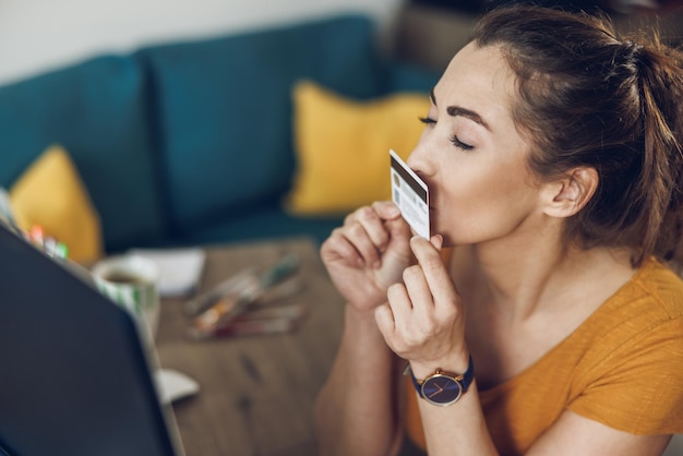 Creative young woman kissing her credit card while using a computer at home