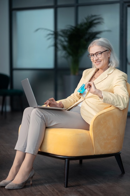 Кредитная карта. Улыбающаяся седая женщина в стильной одежде сидит в удобном желтом кресле с ноутбуком и дебетовой картой
