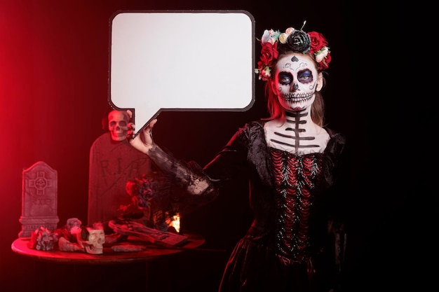 Creepy goddess of death holding speech bubble with sugar skull and flowers crown, having mysterious look. Woman using copyspace template on cardboard, santa muerte advertisement in studio.