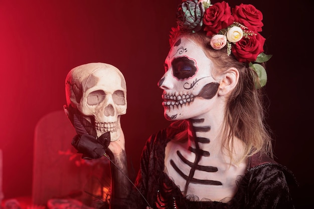 Creepy goddess holding skull and black rose in studio, acting horror like la cavalera catrina in halloween costume. Lady of death celebrating mexican holiday tradition, day of the dead.