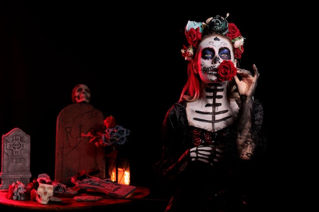 Creepy lady of death holding rose and wearing beautiful skull make up or body art, looking like la cavalera catrina in studio. Glamour woman with santa muerte halloween costume on day of the dead.
