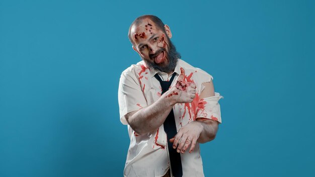 Creepy looking zombie showing thumbs up hand gesture on blue background. Approving spooky brain-eating evil walking dead corpse with deep and bloody wounds showing approval hand sign. Studio shot