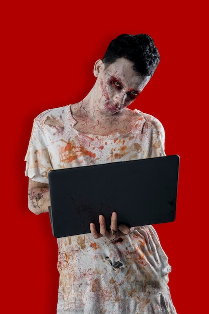 Foto raccapricciante zombie maschio in possesso di un computer portatile e in piedi in studio isolato su sfondo rosso