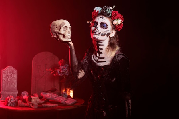 Жуткий молодой человек держит череп в студии, ведет себя страшно и ужасно, чтобы отпраздновать мексиканский хэллоуин. Красивая женщина в праздничном костюме с боди-артом, похожая на богиню смерти.