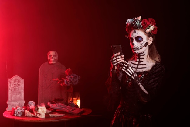 Creepy young person using smartphone over black background, browsing internet website in studio. Santa muerte model portraying horror goddess of death on mexican holiday celebration.
