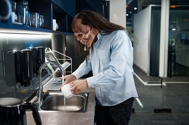 Creepy zombie employee washing cup in sink. Lunch time and coffee break concept. Office dining room interior