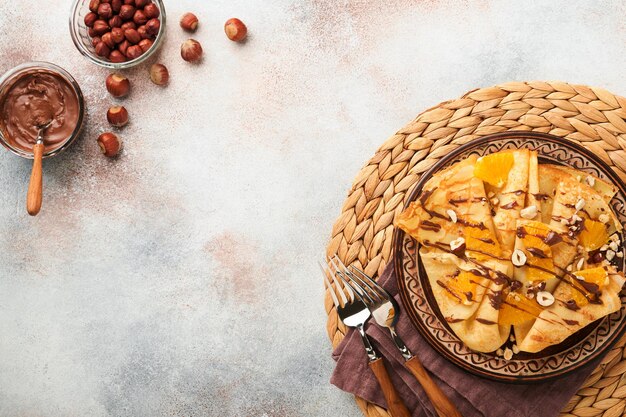 Crepes suzette with oranges and chocolate thin crepes with\
chocolate spread hazelnuts and orange slices fruit in plate for\
breakfast on old concrete rustic table background copy space top\
view